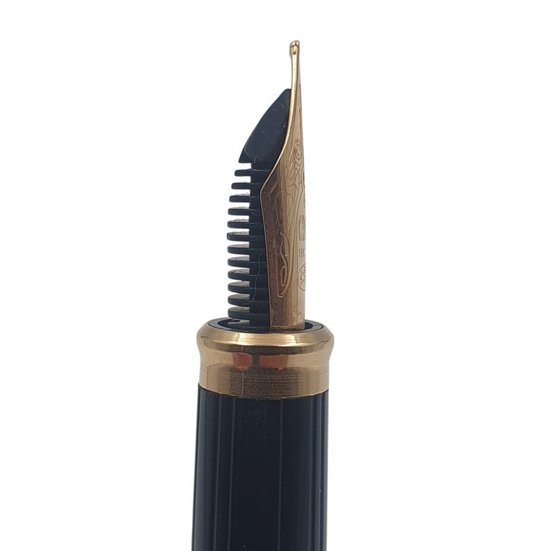 K&ouml;nnte beinhalten: Nahaufnahme einer Feder eines F&uuml;llfederhalters. Die Feder ist goldfarben mit aufwendigen Gravuren und einer schwarzen Zufuhr. Der Stiftk&ouml;rper ist schwarz mit einem goldfarbenen Band in der N&auml;he der Feder.