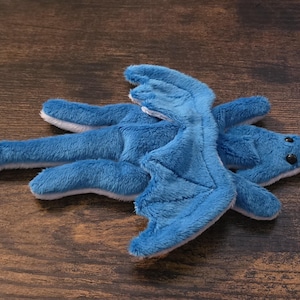 May include: A plush dragon toy made of soft blue fabric with a white underside. The creature features two large wings, a long tail, and two small black plastic eyes. The design is flat and elongated, showcasing a detailed stitched wing pattern.