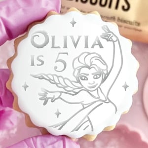 May include: A round scalloped sugar biscuit with white icing featuring a silver-toned graphic of a character with a long braided hairstyle, the text "OLIVIA IS 5", and small four-pointed star accents. The biscuit has a light brown baked edge.
