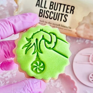 May include: A close-up of a green-iced biscuit with a decorative design, held by a person wearing pink gloves. The biscuit has a scalloped edge and the text "ALL BUTTER BISCUITS" is visible in the background.