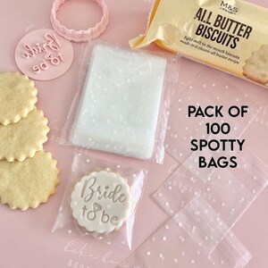 May include: A flat lay featuring wedding-themed baking supplies. Includes a package of biscuits, a cookie cutter, a stamp with "Bride to be", and a stack of cookies. Also, a pack of 100 spotty bags.