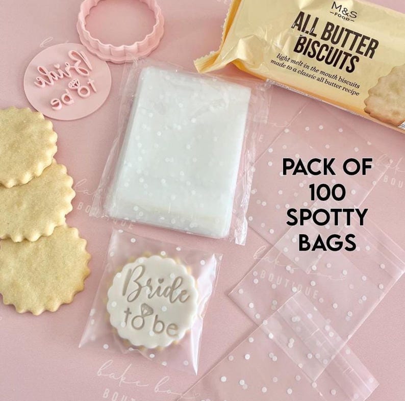 May include: A flat lay featuring wedding-themed baking supplies. Includes a package of biscuits, a cookie cutter, a stamp with "Bride to be", and a stack of cookies. Also, a pack of 100 spotty bags.
