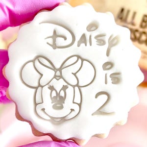 May include: A round, white biscuit with a scalloped edge. The biscuit features a grey embossed image of Minnie Mouse, the text "Daisy is 2", and a bow. The biscuit is held by a person wearing pink gloves.