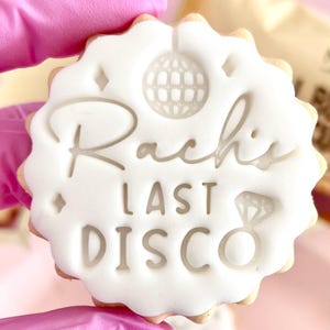 May include: A round cookie with a white icing design. The cookie has a scalloped edge and features the text "Rachi's LAST DISCO" along with a disco ball and a diamond ring illustration.