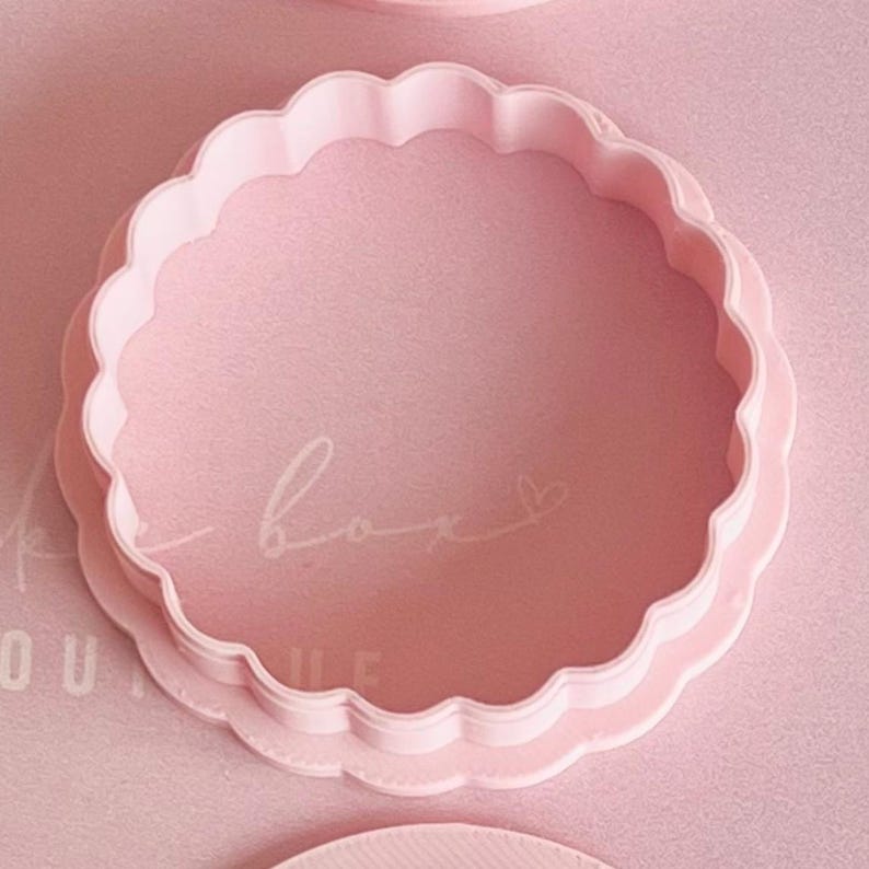 May include: A light pink, scalloped-edge cookie cutter. The cutter has a circular shape with a wavy edge. The image also shows the word "box" in cursive script. The cutter is made of plastic and is likely used for baking.