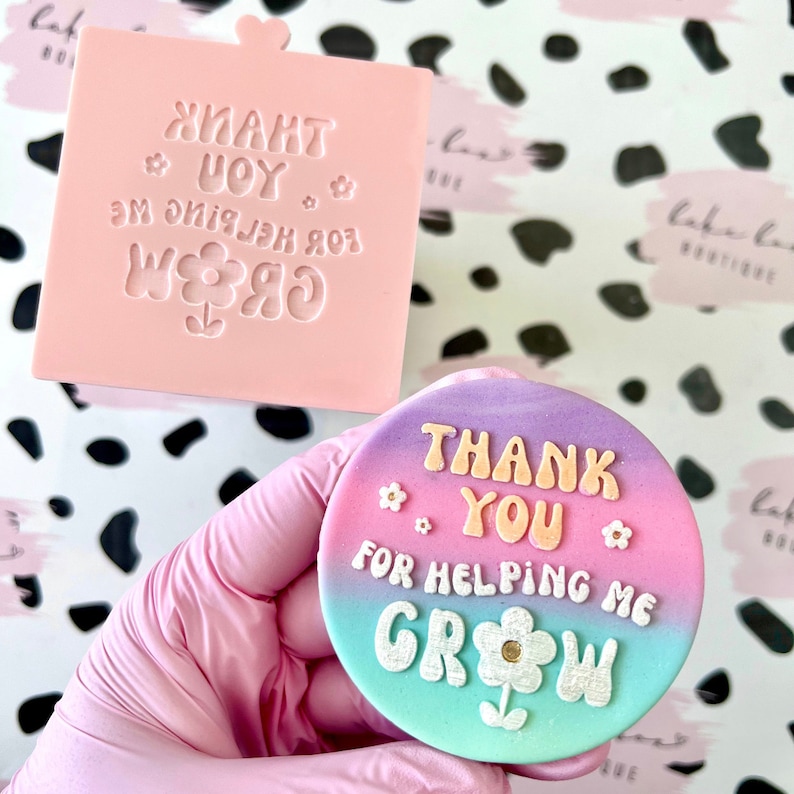 Puede incluir: Un molde de silicona rosa con el texto "Thank You for Helping Me Grow" en una fuente colorida con un dise&ntilde;o de flor. Una galleta decorada con el mismo texto y dise&ntilde;o se muestra en el fondo.