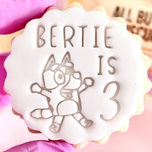 May include: A round, white cookie with a scalloped edge, decorated with the text "BERTIE IS 3" and a cartoon character. The cookie is held by a person wearing pink gloves. A birthday treat.