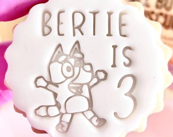 May include: A white cookie with a scalloped edge, featuring the text "BERTIE IS 3" and a cartoon character design. The cookie is held by a person wearing pink gloves. The cookie is a birthday treat.