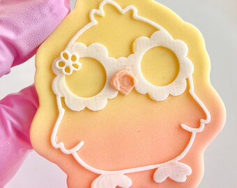 May include: A yellow and orange sugar cookie decorated with white icing in the shape of an owl with large eyes and a flower on its head.