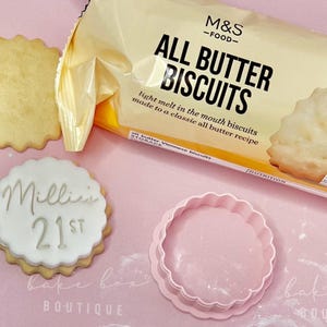 May include: A packet of M&S All Butter Biscuits with a biscuit visible. A decorated biscuit with the text "Millie 21st" is next to a pink cookie cutter. The background is pink.