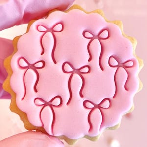 May include: A pink sugar cookie decorated with a repeating pattern of pink bows. The cookie has a scalloped edge.