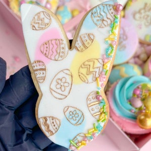 May include: A white bunny-shaped sugar cookie decorated with pastel blue, yellow, and pink icing and gold glitter. The cookie is covered in a pattern of gold-outlined eggs.