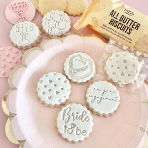 May include: A collection of round, white iced biscuits with various designs, including hearts, bows, and the text "Bride to be" and "on your engagement." The biscuits are arranged on a pink plate with a gold rim, alongside a package of "All Butter Biscuits."