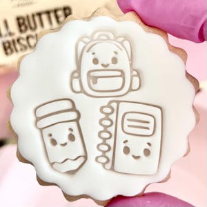 May include: A round biscuit decorated with white icing, depicting a backpack, pencil, and notebook. The biscuit has a scalloped edge and a light brown colour. The design is in a cartoon style, ideal for back-to-school treats.