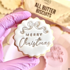 May include: A festive biscuit with white icing, featuring the words "MERRY Christmas" in a decorative font. The biscuit has a scalloped edge and a bow design. The biscuit is held by a gloved hand.