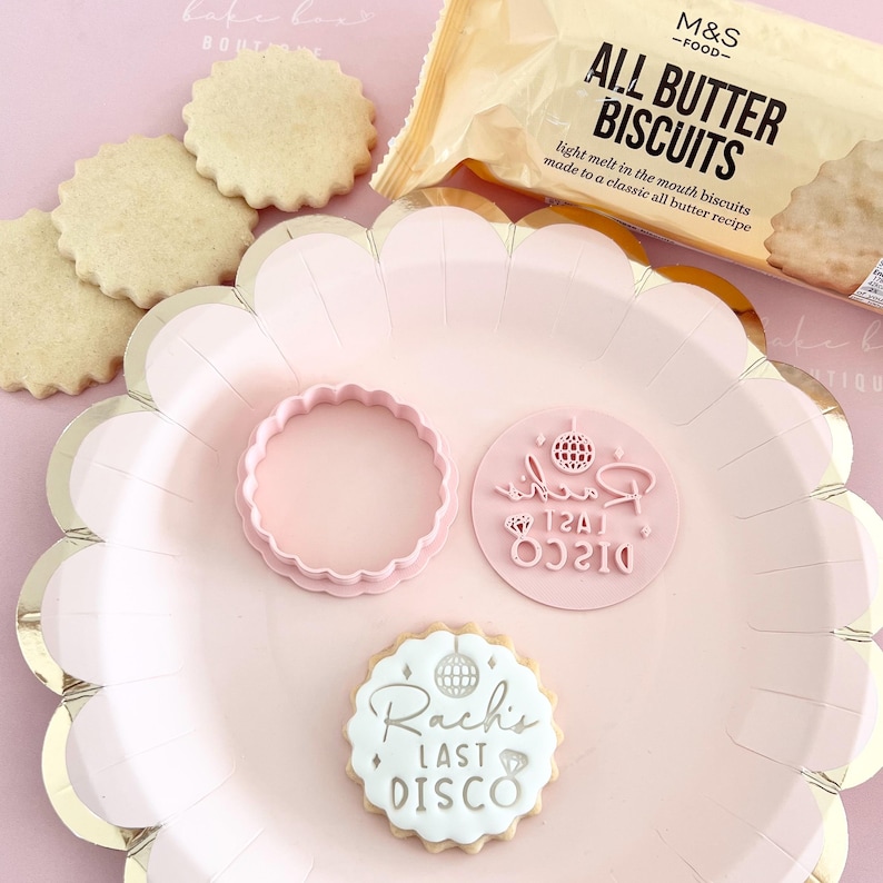 May include: A pink scalloped plate holds a cookie cutter, stamp, and decorated cookie. The cookie is stamped with "Rach's LAST DISCO" and a disco ball. A package of "ALL BUTTER BISCUITS" is in the background. The cookies are light brown.