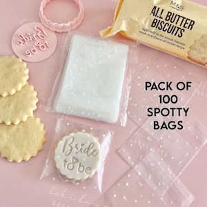 May include: A selection of baking items for a wedding theme. Includes a packet of biscuits, a cookie cutter, a stamp with "Bride to be", and a stack of cookies. Also, a pack of 100 spotty bags.