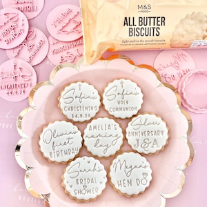 May include: A pink plate holds decorated biscuits with white icing and silver lettering for special occasions. The biscuits are labeled for events like christenings, birthdays, and bridal showers. Pink cookie stamps and a package of all butter biscuits are also visible.