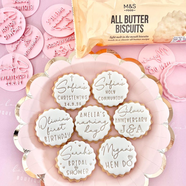 May include: A pink plate holds decorated biscuits with white icing and silver lettering for special occasions. The biscuits are labeled for events like christenings, birthdays, and bridal showers. Pink cookie stamps and a package of all butter biscuits are also visible.