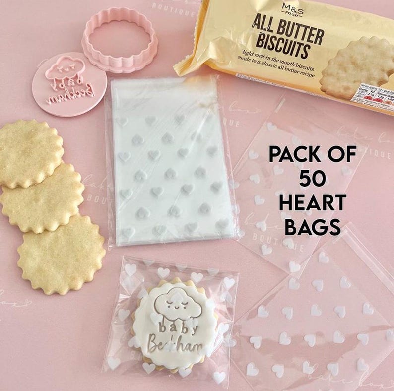 May include: A collection of baking supplies and treats. Includes a package of biscuits, heart-shaped bags, a cookie cutter, and cookies. One cookie is decorated with a cloud and the text "baby Beckham". The bags are labeled "PACK OF 50 HEART BAGS".