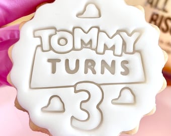 Può includere: Un biscotto rotondo con glassa bianca. La glassa presenta le parole "TOMMY TURNS 3" in grassetto. Il biscotto ha un bordo smerlato ed è tenuto da una persona con guanti rosa. Una dolcezza per il compleanno.