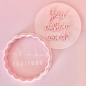 May include: A light pink cookie cutter set. One cutter is round with the words "Your custom words" in a script font. The other is a scalloped circle with the word "Boutique".