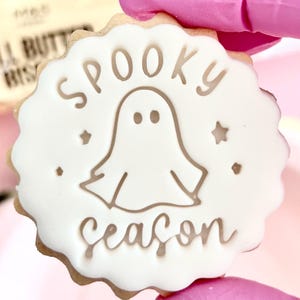 May include: A round biscuit with a scalloped edge, decorated with white icing. The icing features a ghost illustration and the words "Spooky Season" in a playful font. Small star and dot accents surround the ghost.