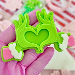 May include: A green cookie cutter shaped like a heart with arms and a crown. The arms have pink cuffs with white trim. The cookie cutter is held by a person wearing a pink glove. Other cookie cutters are in the background.