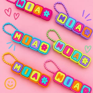 May include: Six colourful keychains featuring the name MIA in block letters followed by a flower icon. Each keychain consists of four rectangular segments in various bright colours, attached to a matching ball chain for easy hanging.