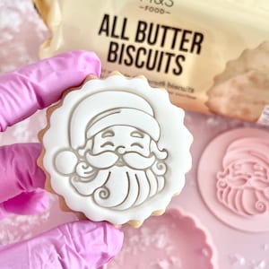 May include: A festive biscuit decorated with a white Santa Claus design. The biscuit is held up, showing its scalloped edge. A pink stamp and a packet of "All Butter Biscuits" are visible in the background.