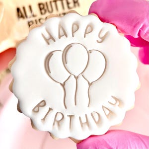May include: A round biscuit decorated with white icing. The icing features the words "HAPPY BIRTHDAY" and three balloon illustrations. The biscuit is held by a person wearing a pink glove.