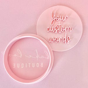 May include: Two pink, circular custom stampers with the words "Your custom words" and "Boutique Lad" in a cursive font. The stampers are on a pink background. The stampers are likely used for crafting or branding.