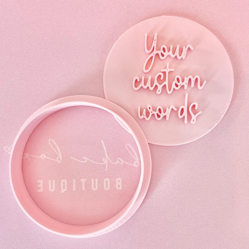 May include: Two pink, circular custom stampers with the words "Your custom words" and "Boutique Lad" in a cursive font. The stampers are on a pink background. The stampers are likely used for crafting or branding.