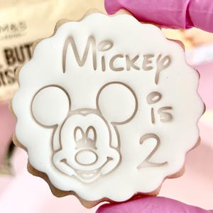 May include: A round biscuit with a white icing design featuring Mickey Mouse's face and the words "Mickey is 2". The biscuit has a scalloped edge and a light brown base.