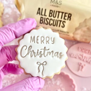 May include: A festive biscuit with white icing, featuring the words "MERRY Christmas" and a bow design. The biscuit has a scalloped edge and is held by a person wearing a pink glove. A packet of "ALL BUTTER BISCUITS" is visible in the background.