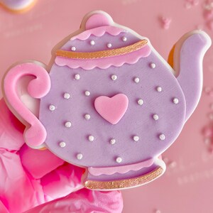 May include: Purple teapot cookie decorated with white dots and a pink heart. The cookie has a gold trim around the top and bottom.