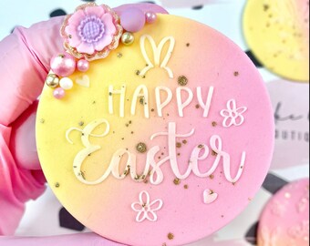 May include: A round, pastel-colored Easter treat with a gradient of yellow to pink. The words "Happy Easter" are written in white script, with bunny ears and floral decorations. Gold sprinkles and decorative elements add detail.
