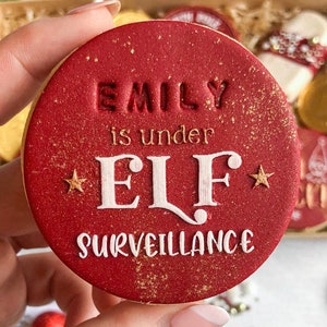 May include: Red Christmas cookie with gold glitter and white icing. The cookie says "Emily is under Elf Surveillance".