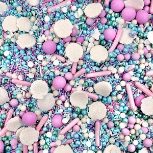 May include: A close-up of a mix of colourful sprinkles. The mix includes light blue, pink, white, and purple sprinkles in various shapes and sizes, including small balls, rods, and star shapes. The sprinkles are scattered across the surface.