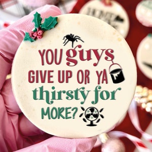 May include: White round bauble with green holly leaves and red berries. The bauble has a black spider, a red paint can, and a green fan with the text "YOU guys GIVE UP OR YA thirsty for MORE?"