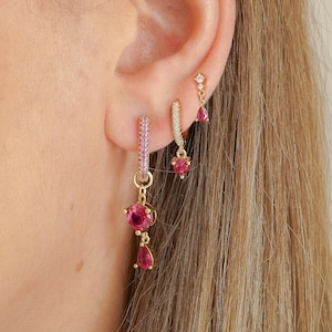 May include: Three gold earrings worn in a single ear. The lowest earring is a hoop with a pink stone drop. The middle earring is a small hoop with a pink stone charm. The highest earring is a small stud with a tiny pink teardrop stone.