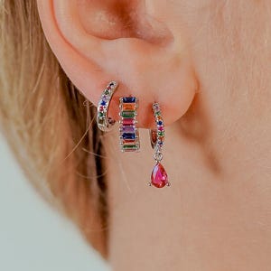 May include: Three pairs of earrings are shown on an ear. The first is a small silver hoop with colourful gemstones. The second is a silver hoop with a rectangular rainbow gemstone design. The third is a silver hoop with a pink teardrop gemstone.