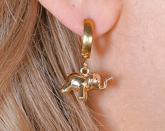 May include: A gold-colored hoop earring with a small, detailed elephant charm. The elephant charm is suspended from the hoop, showcasing the jewelry's design. The earring is worn on a person's ear, with their blonde hair visible in the background.