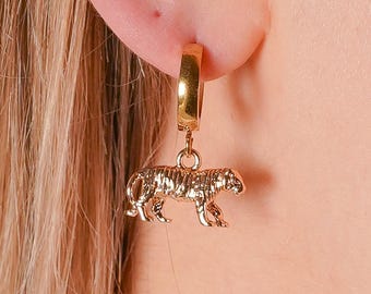 May include: Gold-colored hoop earring with a small, detailed tiger pendant. The tiger is in a walking pose, with stripes visible. The earring is attached to an ear, with blonde hair in the background. The jewelry is a fashion accessory.