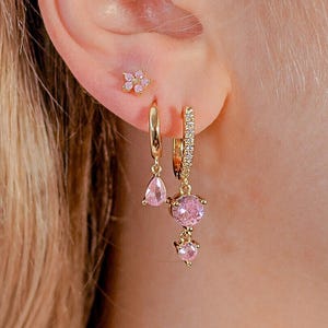 May include: A close-up of an ear displaying three pieces of jewellery. A small, gold flower stud is in the upper ear. Two gold hoop earrings with pink gemstones hang below. One earring has a teardrop gem, the other has a round gem and a smaller gem.