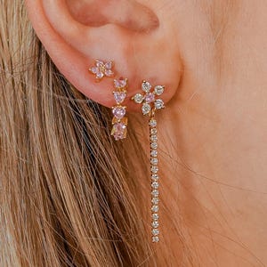 Op de afbeelding: Close-up van drie goudkleurige oorbellen op een oor. De oorbellen hebben roze en heldere edelstenen in bloem- en hartvormen. Eén oorbel heeft een lang, bungelend ontwerp.