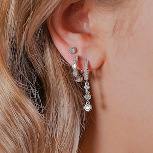 Puede incluir: Primer plano de una oreja adornada con múltiples pendientes plateados. Los pendientes incluyen un pequeño perno, un par colgante con múltiples piedras transparentes y un pendiente de aro con piedras transparentes. Los pendientes brillan sobre el cabello rubio.