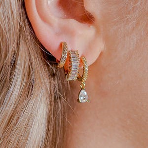 May include: Gold-coloured hoop earrings featuring clear, rectangular and pear-shaped cubic zirconia stones. The earrings are displayed on an ear, highlighting the jewellery's design and sparkle. The earrings combine classic and modern styles.
