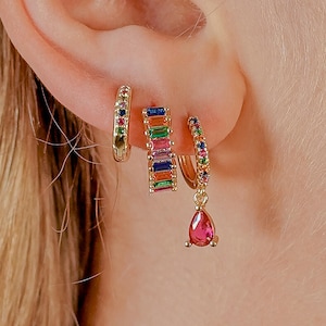 Puede incluir: Juego de pendientes dorados. Un par es un aro pequeño con gemas de colores. Otro tiene forma rectangular con gemas de colores. El tercero es un aro con un colgante de gema rosa en forma de lágrima.