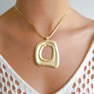 May include: A gold chain necklace with a large, abstract, gold pendant. The pendant is a double-layered, irregular square shape with a smooth, hammered finish.